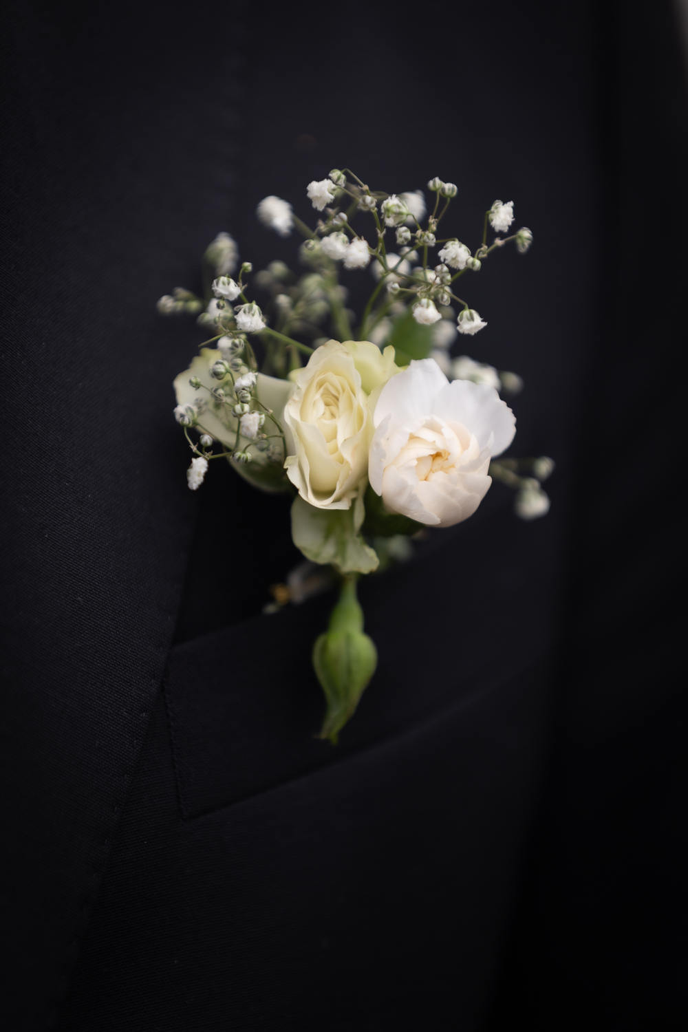 Nærbilde av brudeknappehull med hvit blomst mot mørk dress, bryllupsfoto av Prospectxmedia Norge.