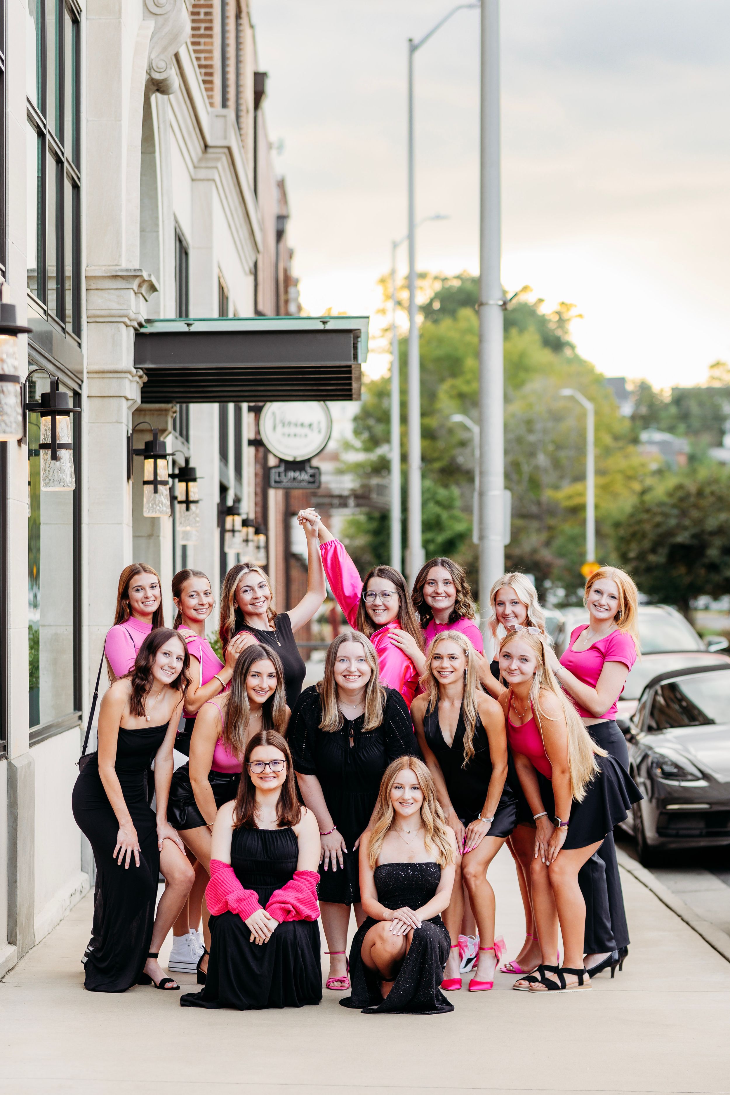 Group of women posing in pink and black outfits on urban sidewalk at sunset.