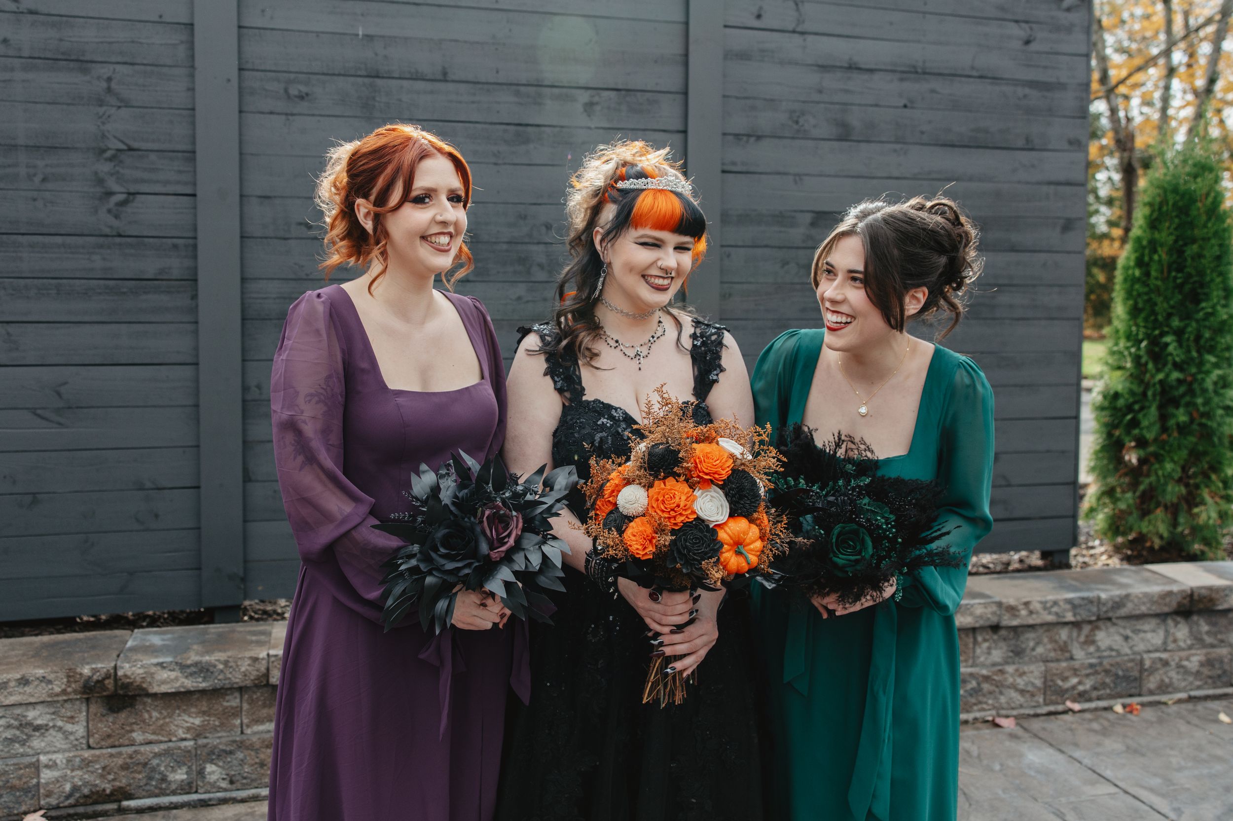 Wedding party members in purple and green dresses hold vibrant orange and dark bouquets against a gray wall.