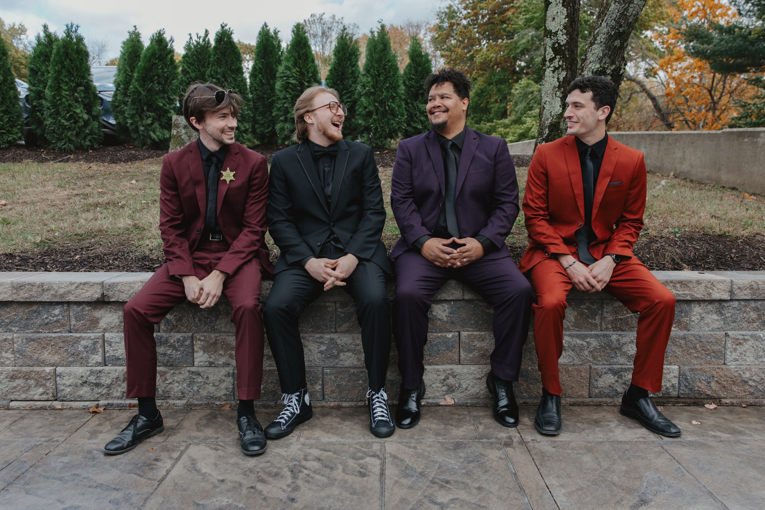 Four people sit together on a stone wall wearing coordinated burgundy and orange outfits in an autumn setting.
