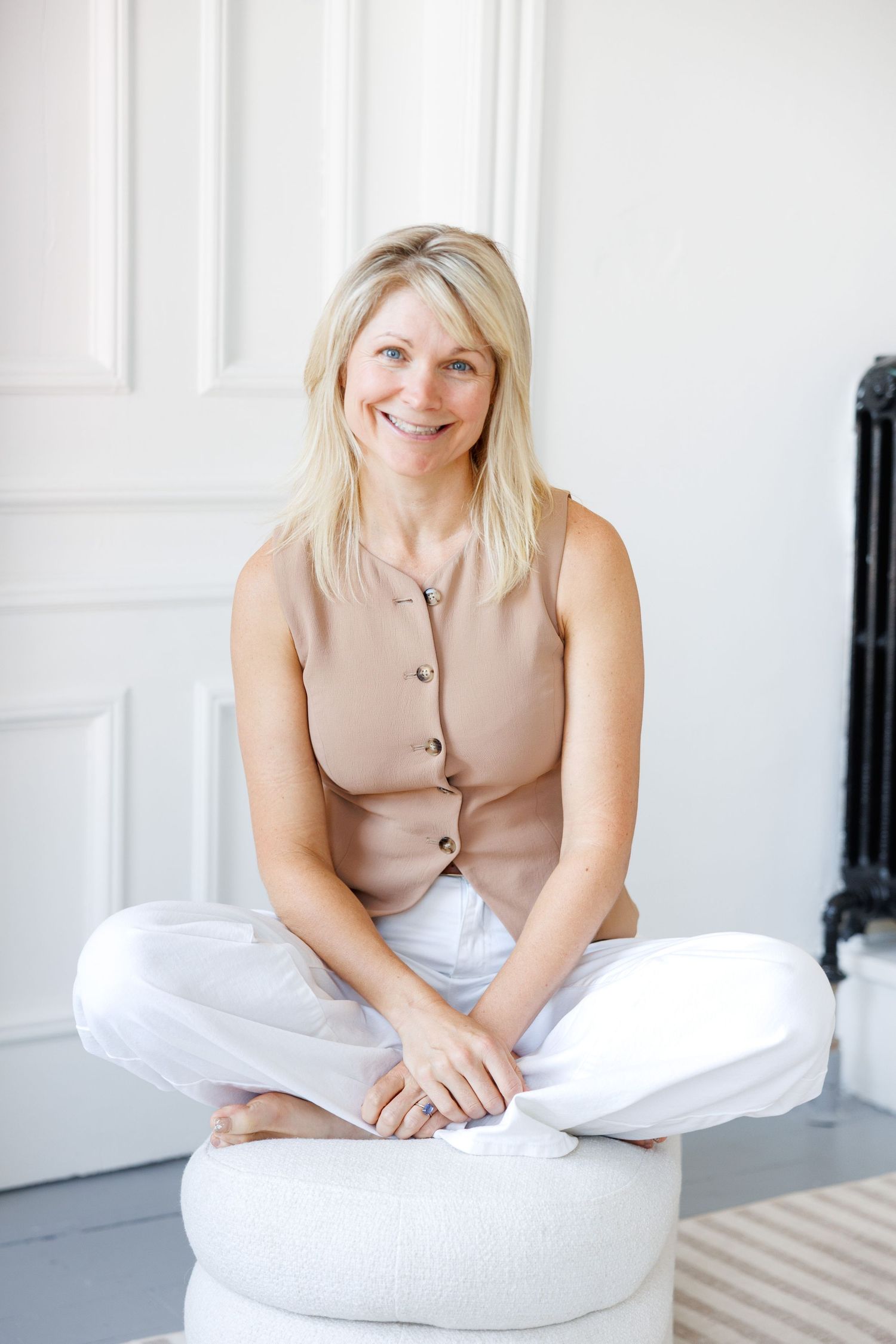 Woman in beige sleeveless top relaxes on white cushions in bright modern interior setting.