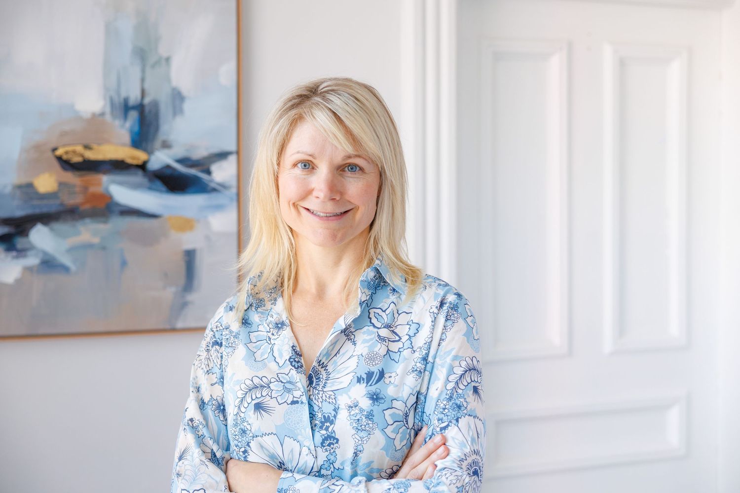 Woman in blue and white floral dress smiles while sitting on white chair in bright modern room.