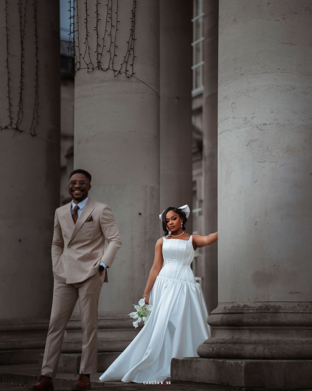 A couple in wedding attire poses joyfully between elegant white columns of a classic building.