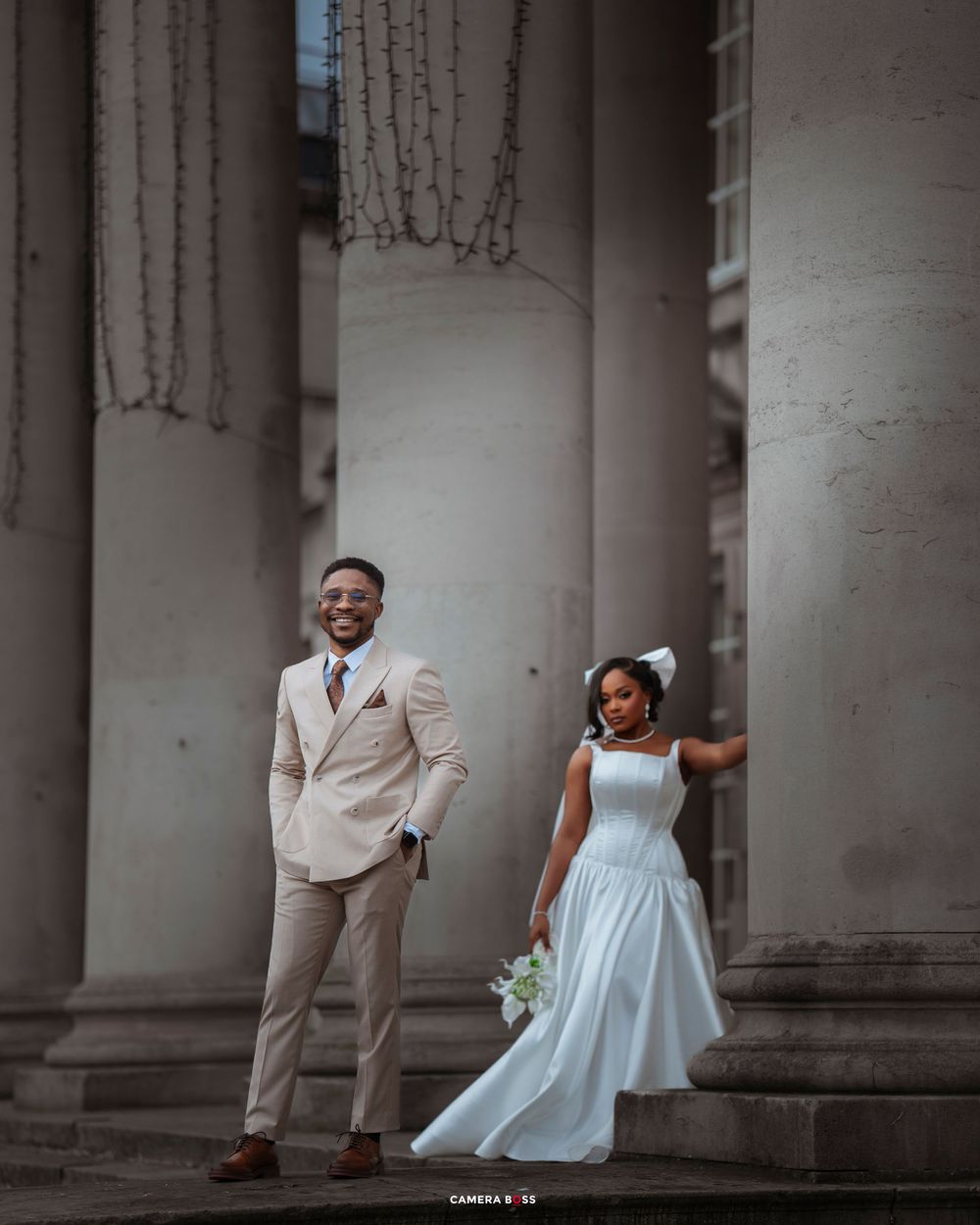 A couple in formal attire shares playful moments between elegant stone columns during a romantic photoshoot.