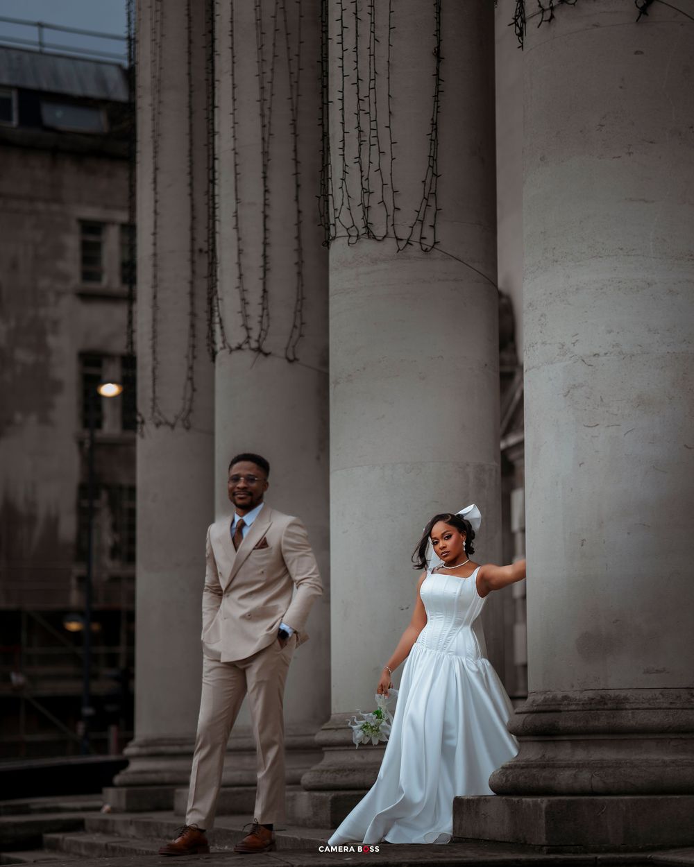 A couple poses elegantly between white classical columns at dusk, with the bride in a flowing white dress and groom in beige suit.