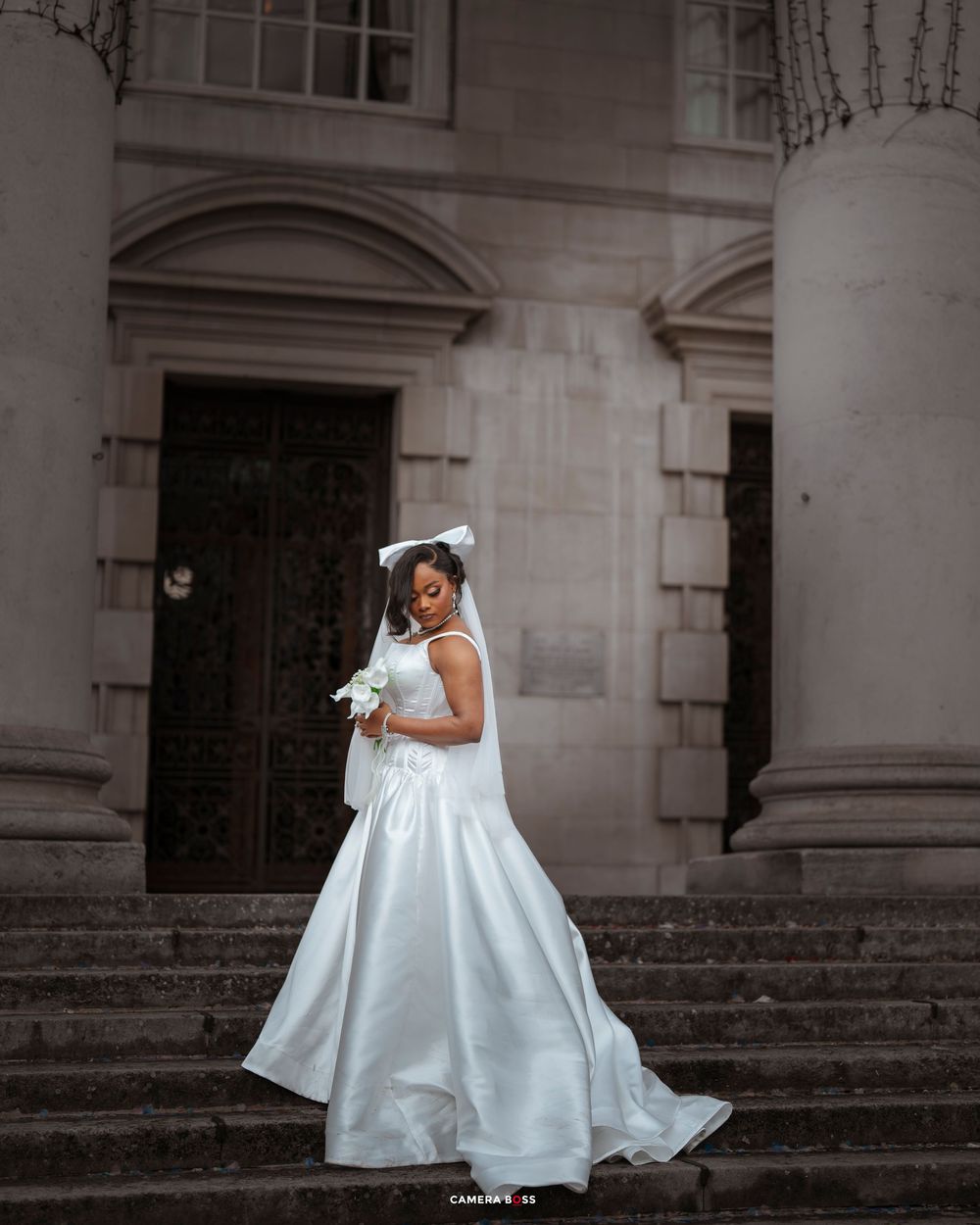 A bride in a flowing white satin wedding gown poses elegantly on stone steps of a historic building with columns.