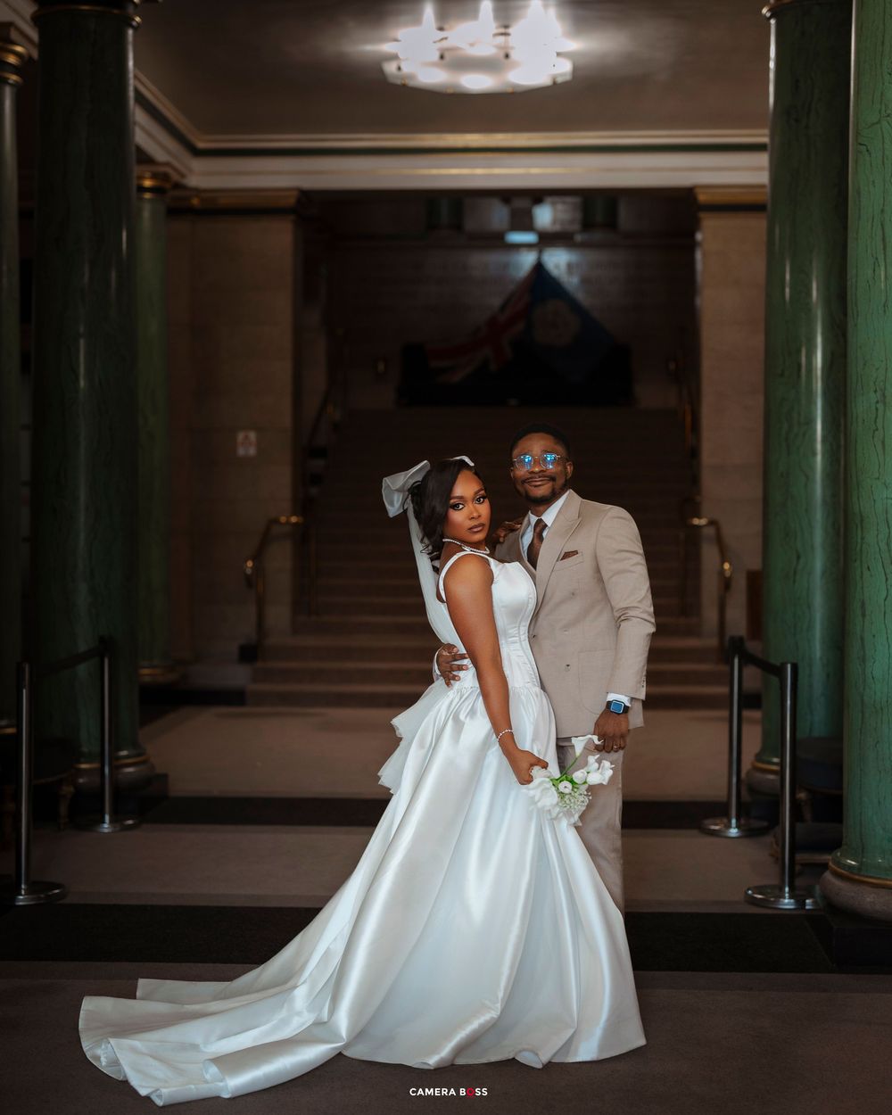 A couple poses elegantly in formal wedding attire inside an ornate hallway with green columns and chandelier lighting.