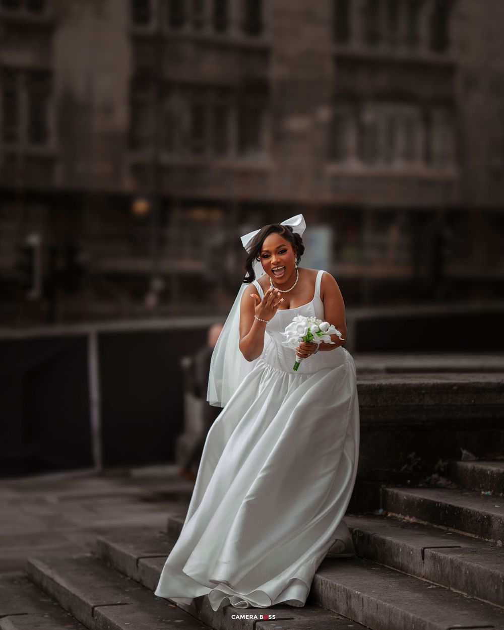 A bride in a flowing white dress sits elegantly on concrete steps in a moody urban setting.