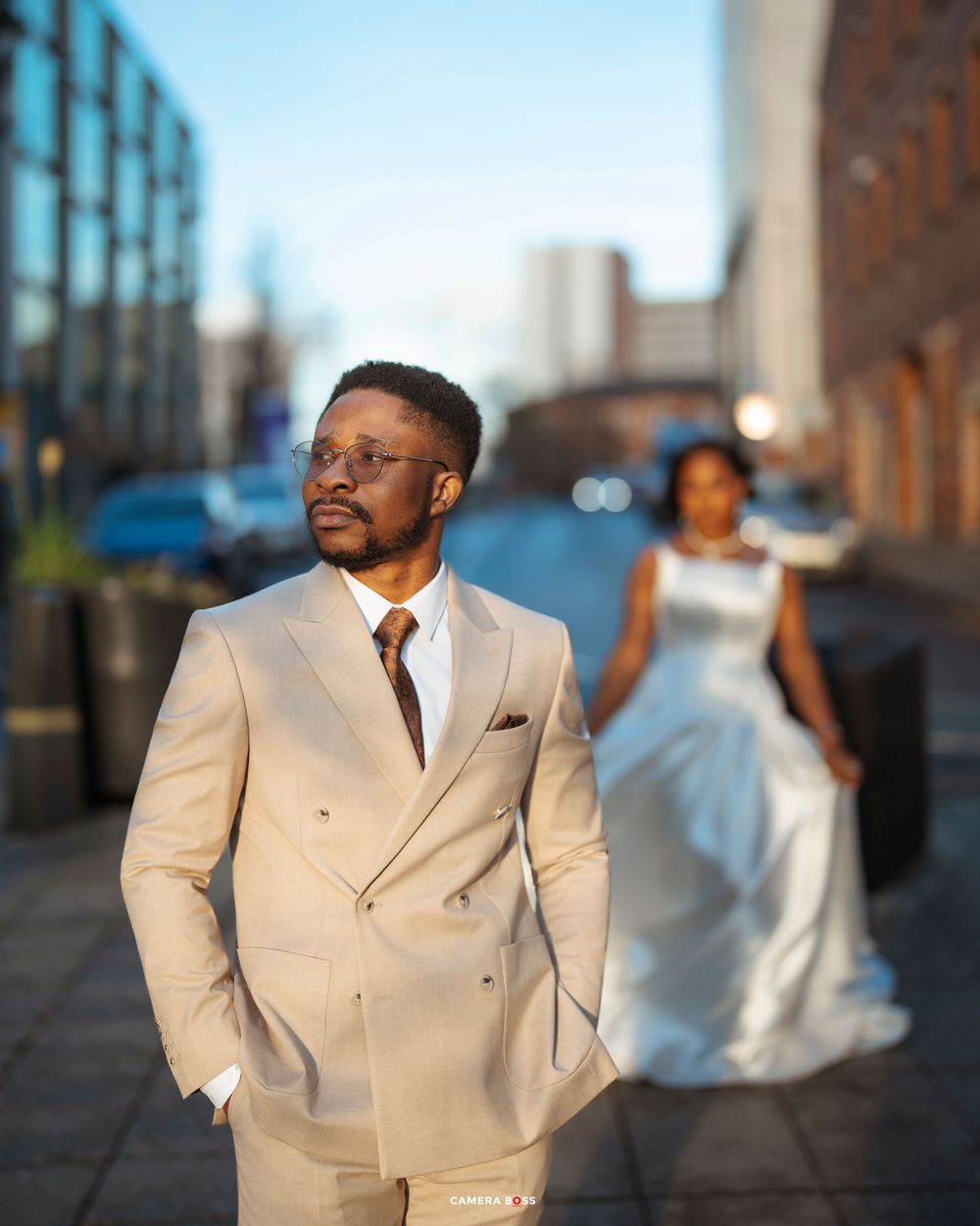 Professional portrait of a person in a cream-colored suit jacket posing against a blurred urban background at sunset.