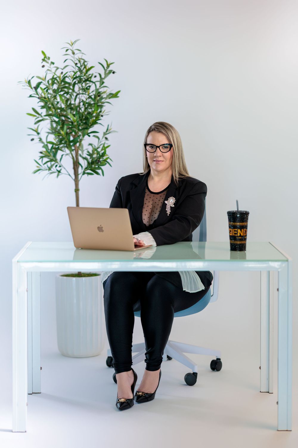 Professional blonde woman in black blazer working on MacBook laptop at white glass desk in office.