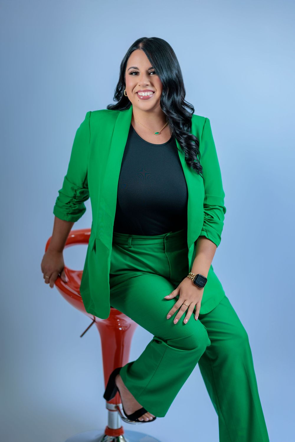 Woman in green blazer and pants suit smiling while seated on red stool against light blue background.