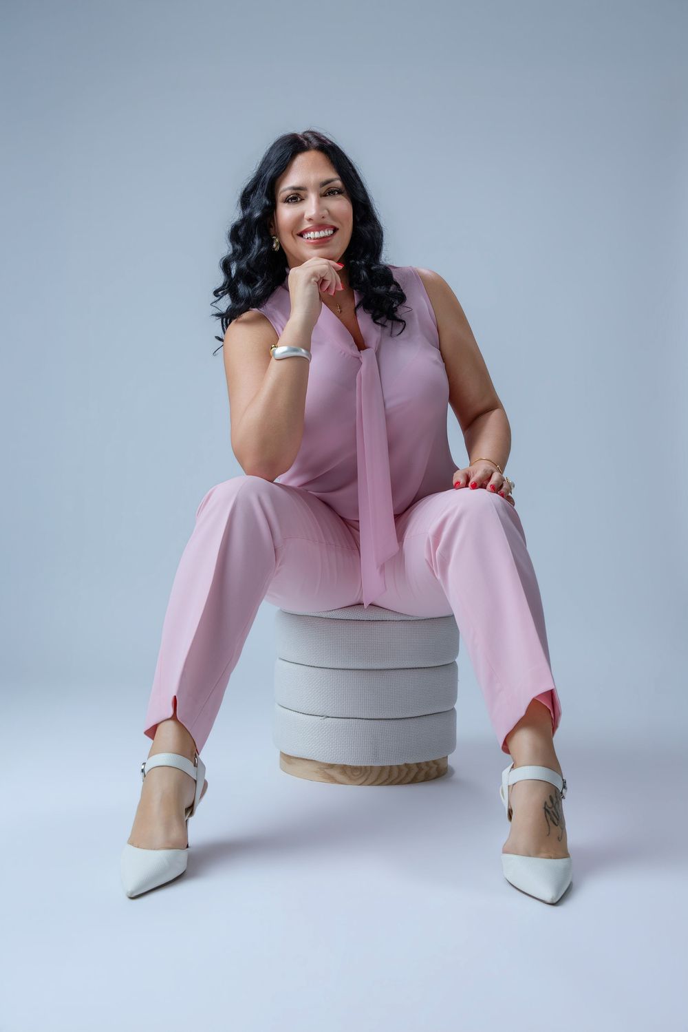Woman in pink jumpsuit sitting on round stool, smiling confidently, with white heels and dark curly hair.