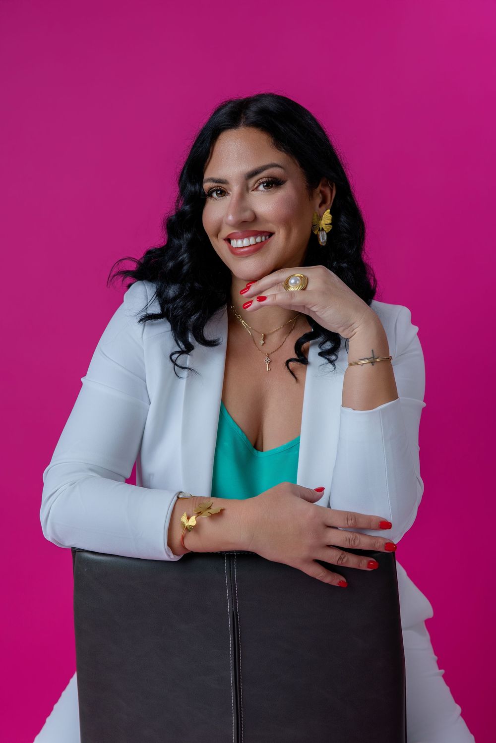 Smiling woman in white blazer and teal top poses against vibrant pink background, wearing gold jewelry and red nails.