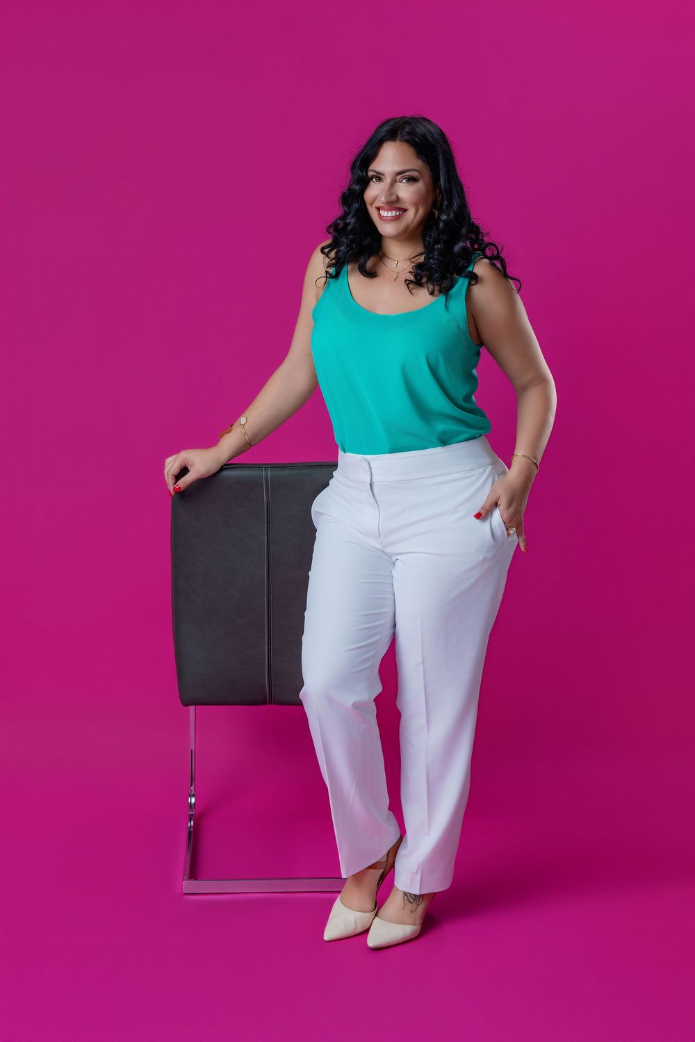 Smiling woman in teal top and white pants leaning on a chair against a vibrant pink background.