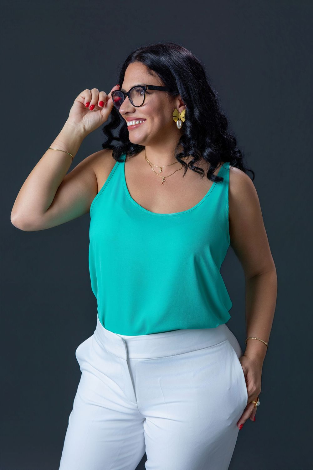Smiling woman in teal tank top and white pants adjusting glasses, with curly black hair and gold earrings.