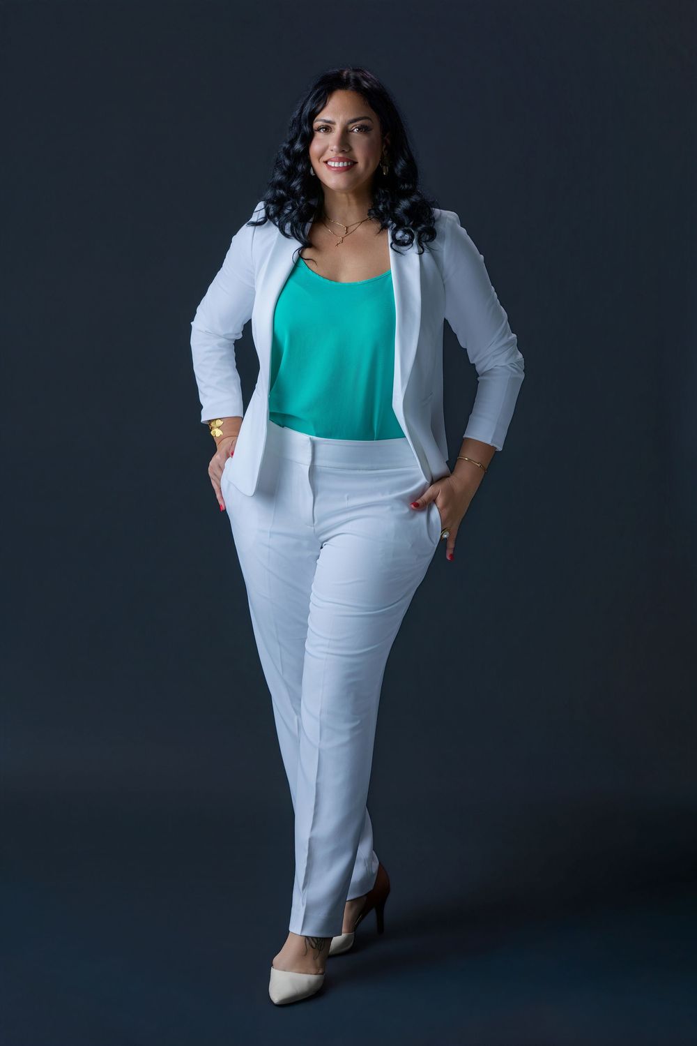 Woman in white suit and teal top posing confidently against dark background.