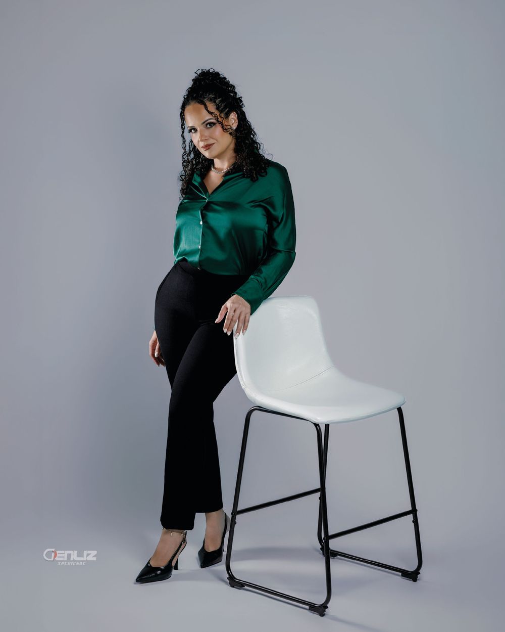 Woman in green satin blouse and black pants posing beside a white chair in a professional studio setting.