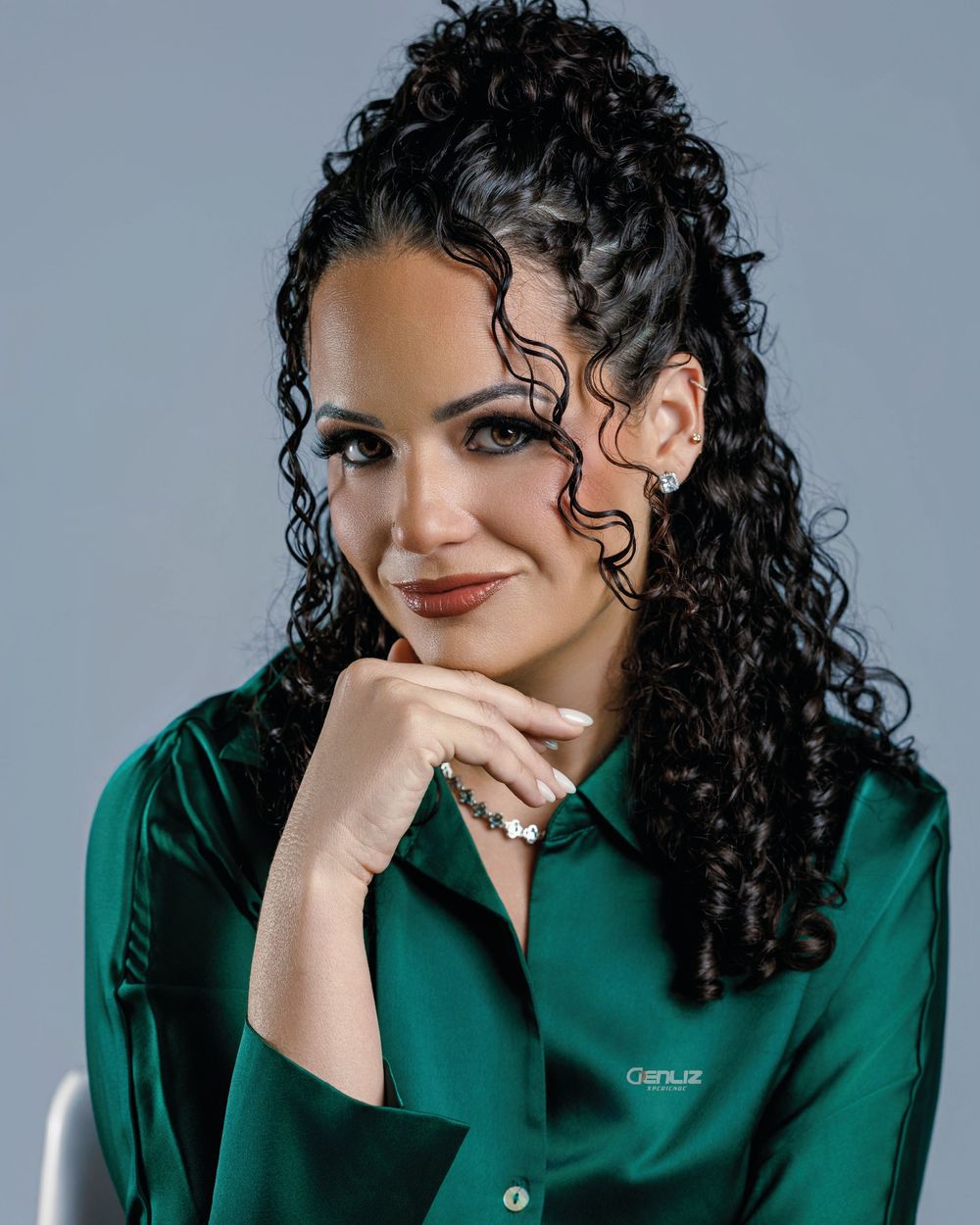 Woman with curly black hair smiling in a green satin shirt, posing confidently with hand on chin.
