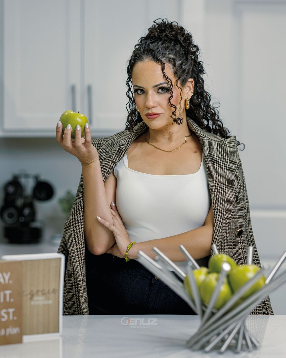 Woman in plaid blazer holding a green apple in a modern white kitchen with fresh apples on the counter.
