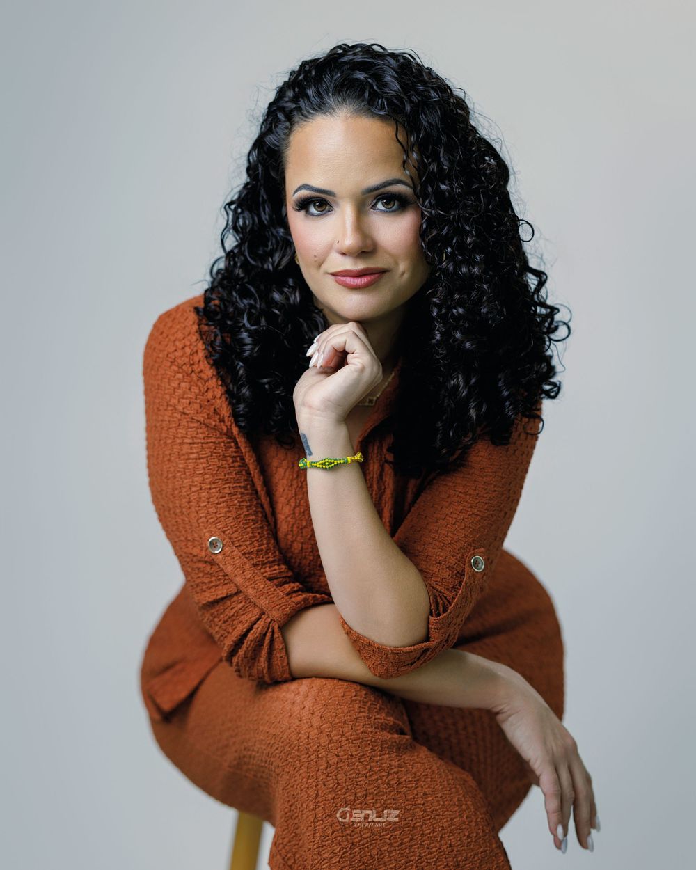 Woman with curly black hair in rust orange knit dress poses gracefully on stool in professional studio portrait.