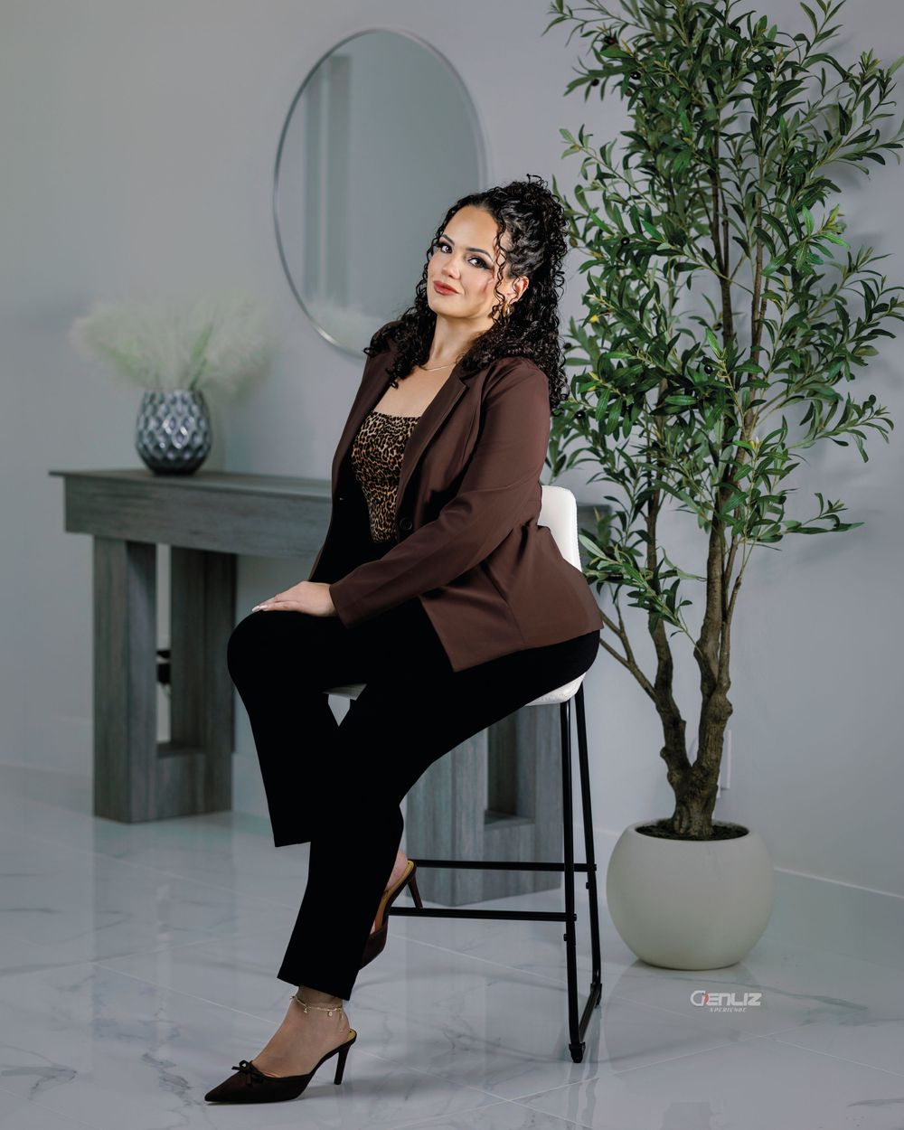 Woman in brown blazer and black pants sits elegantly on a stool beside a green plant in a modern white studio.