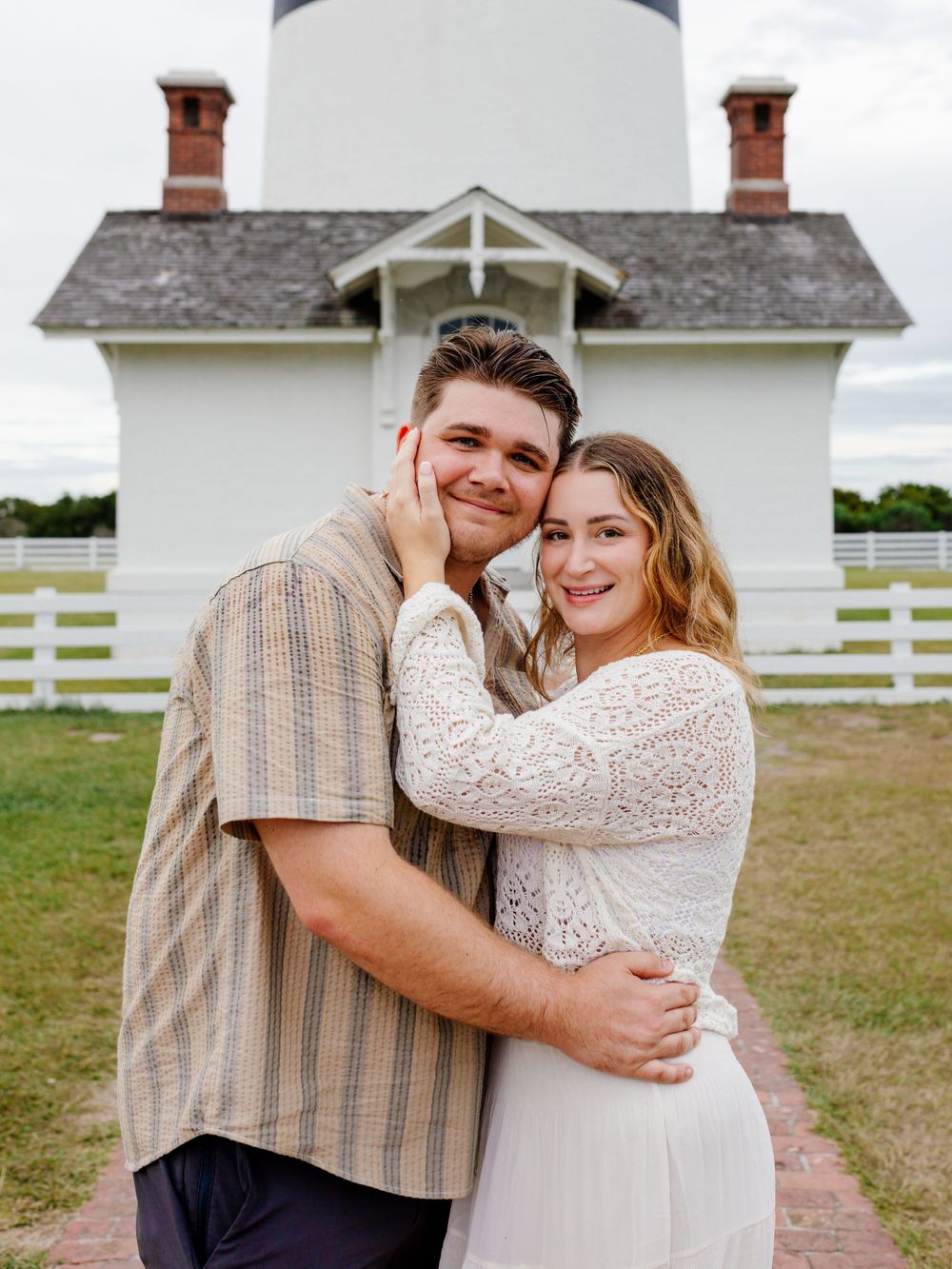 A couple embraces in front of a lighthouse on a cloudy day.