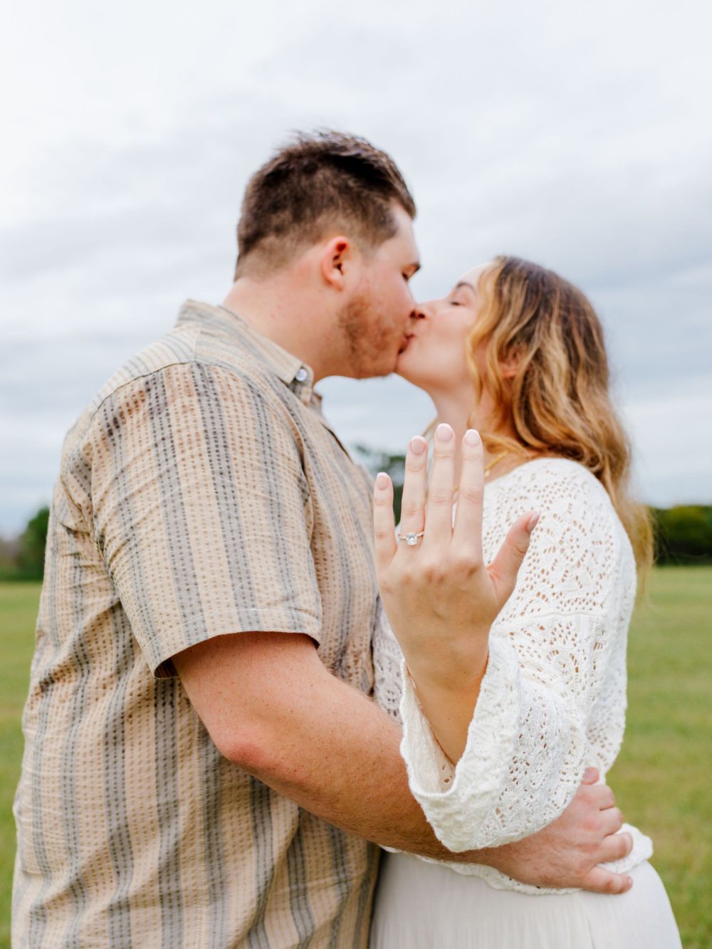 A romantic couple embraces in a grassy field wearing coordinated beige and white outfits.