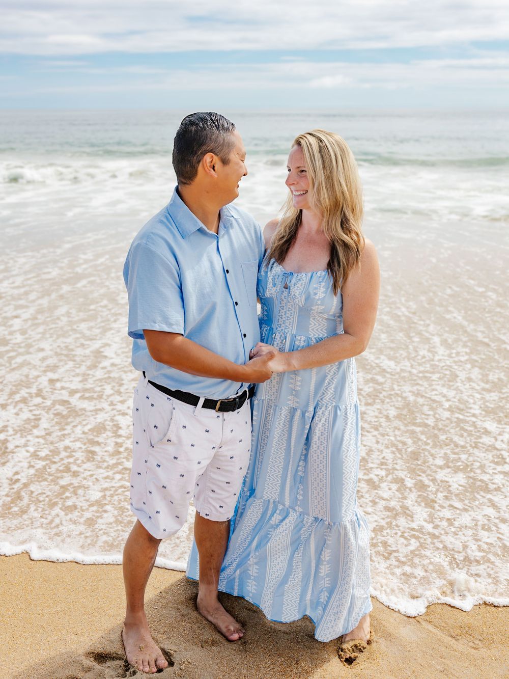 A couple shares loving moments on a beach with waves rolling in behind them.