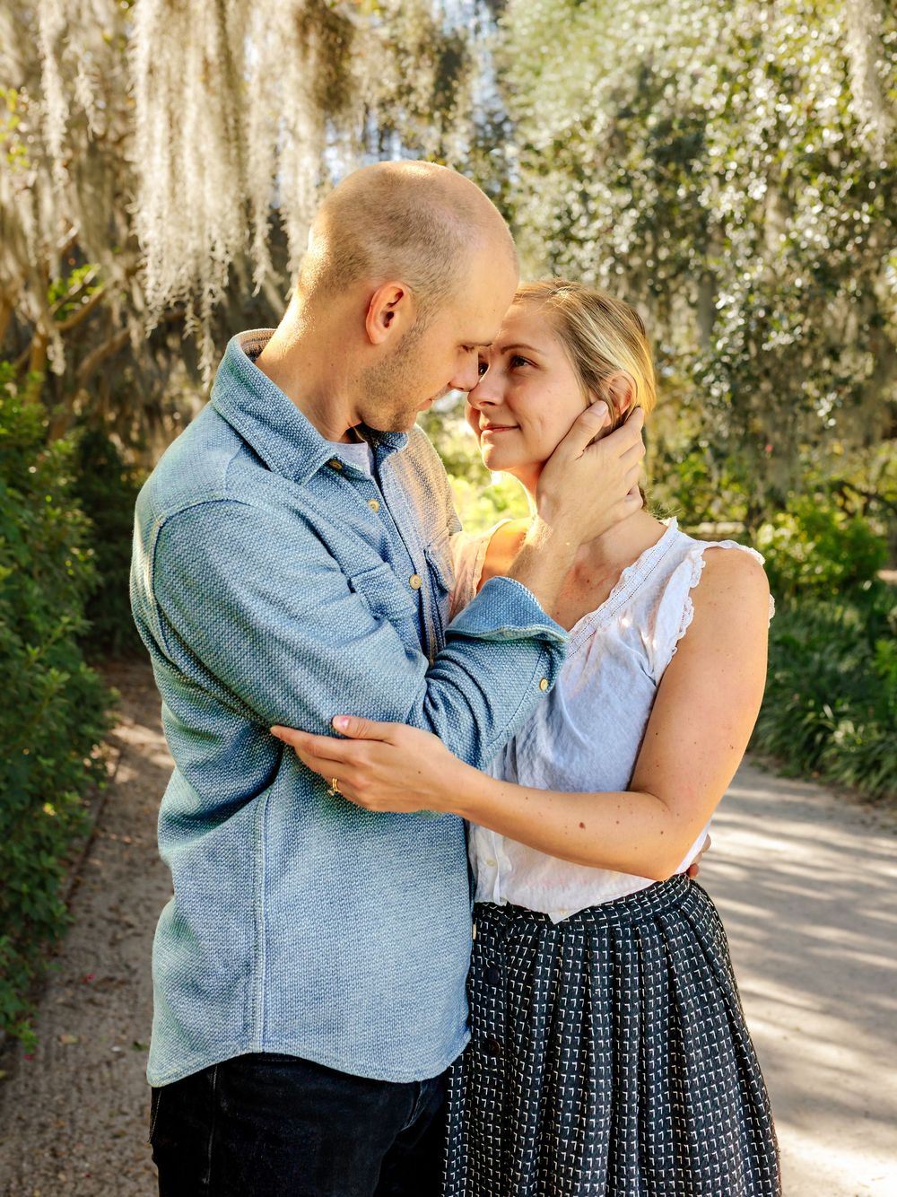 A romantic couple embraces on a sunlit path beneath Spanish moss draped trees.