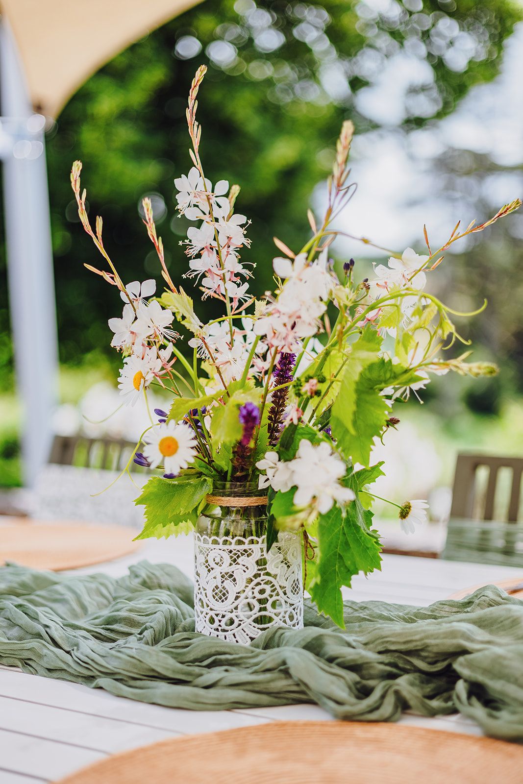 Elegant floral centerpiece with white flowers and greenery in lace-wrapped vase on sage green table runner.