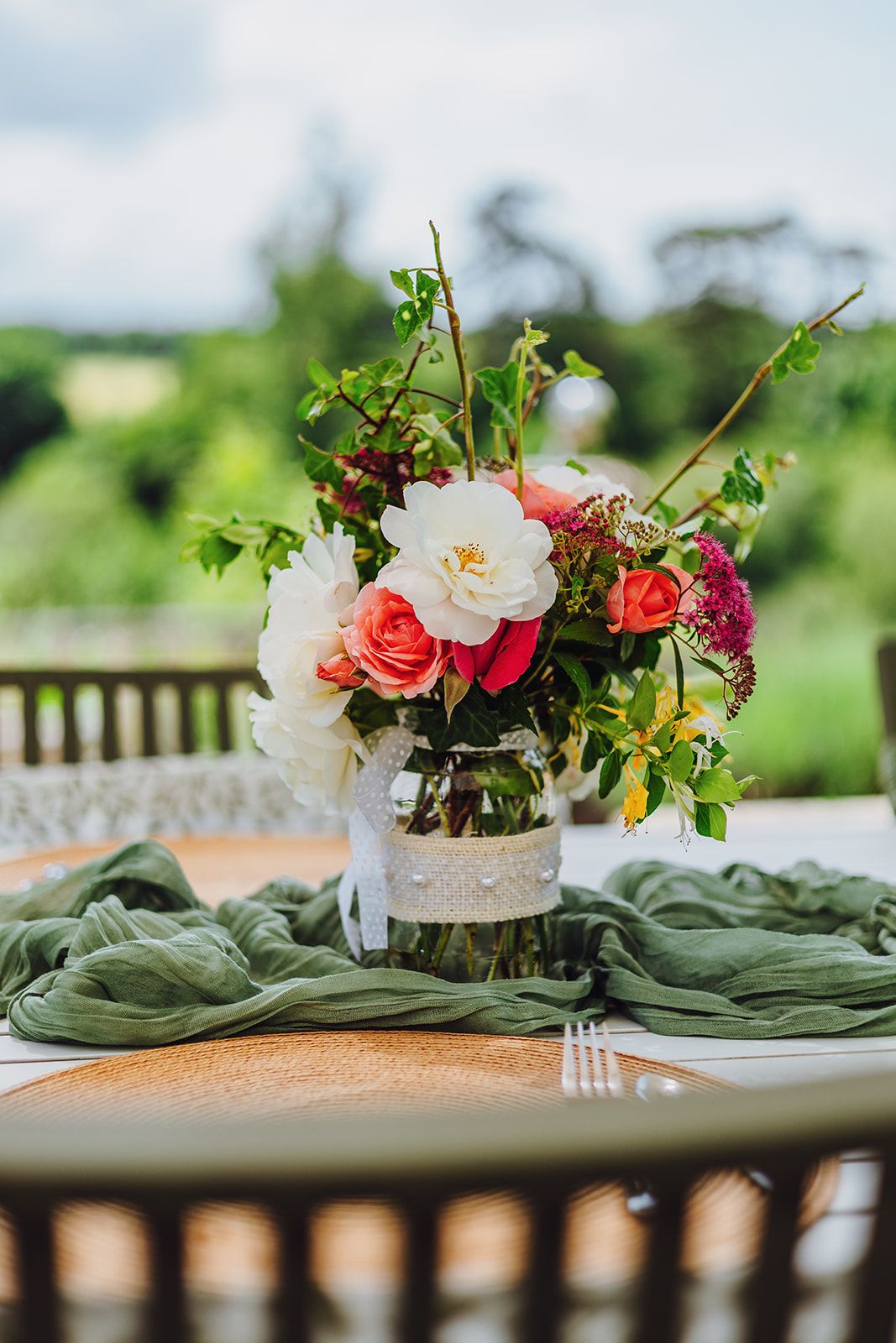 Elegant outdoor table setting with coral and white floral arrangements in lace-wrapped vases and sage green napkins.