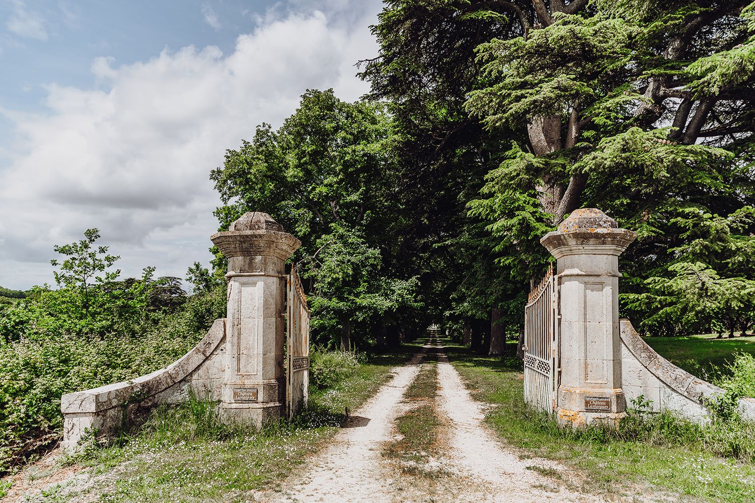 A rustic stone entrance gate with pillars leads down a dirt path through green trees and foliage.