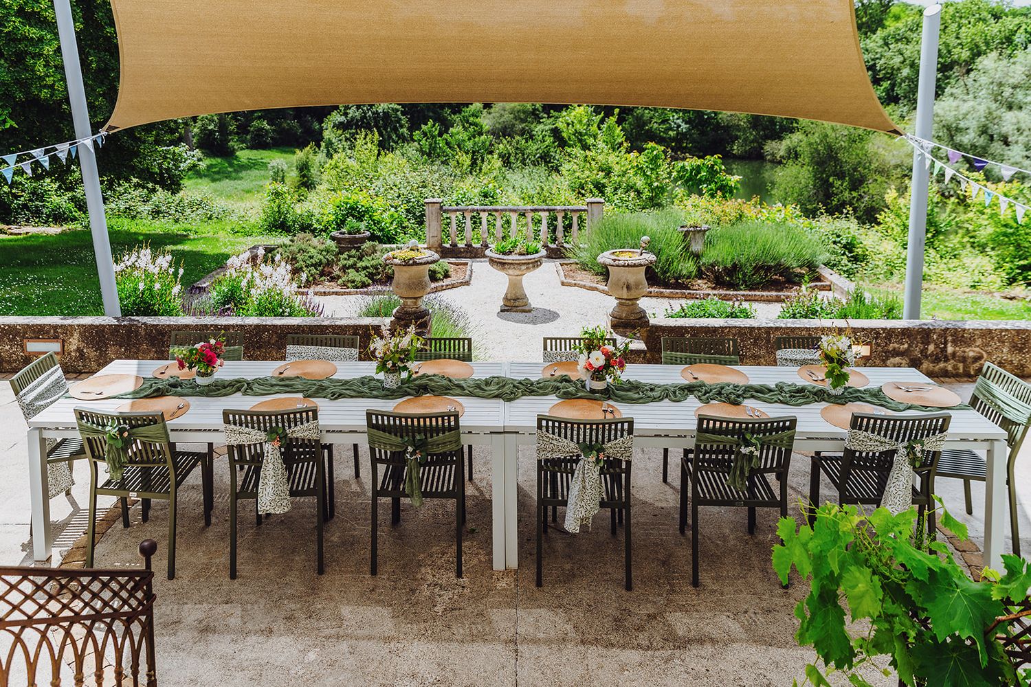 Outdoor restaurant patio with wooden tables and chairs under a beige canopy surrounded by lush greenery.