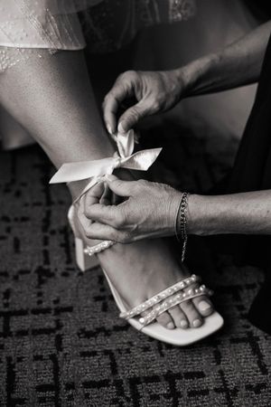 Black and white photo of hands tying ribbon bow on bride's pearl-strapped wedding heels before ceremony.