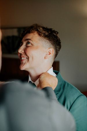 Smiling person in green sweater getting tie adjusted, looking up joyfully during wedding preparations.