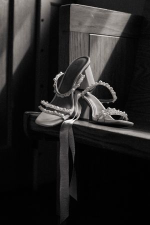 Black and white photo of elegant pearl-adorned bridal heels with ribbon ties resting on a wooden chair.