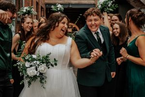 Two brides laughing and holding hands walking through cheering wedding guests in green dresses.