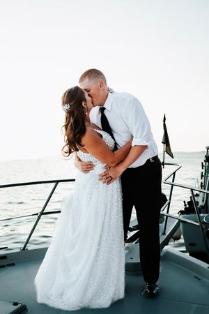 Bride and groom sharing a romantic kiss on the bow of a boat at sunset over water.