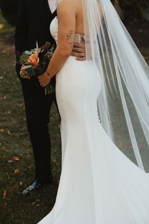 Bride in fitted backless wedding gown with cathedral veil held by groom in black suit outdoors.