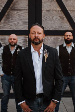 Groom in black blazer stands confidently before groomsmen in vests against rustic wooden backdrop.