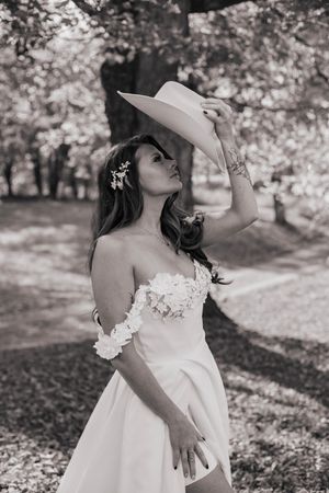 Black and white photo of bride in off-shoulder gown holding wide-brim hat outdoors among trees.