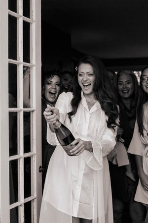 Black and white photo of bride in white robe popping champagne bottle with bridesmaids behind her.