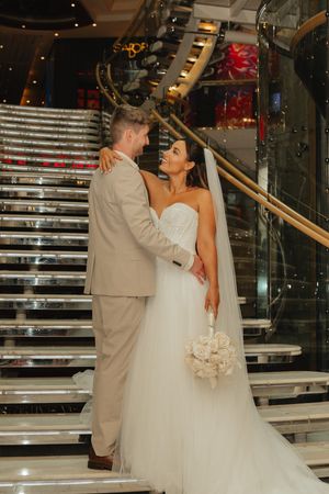 Bride and groom share a romantic moment on a grand staircase, she holds a white bouquet in her wedding gown.