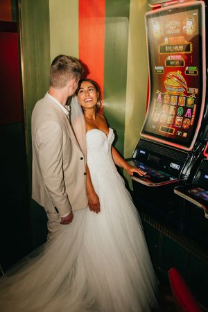 Newlyweds pose together smiling beside a slot machine in a casino, bride in white gown and groom in tan suit.