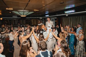 Bride and groom lifted by cheering wedding guests on a crowded dance floor during lively reception celebration.
