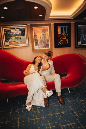 Bride and groom sit closely on a red heart-shaped sofa, laughing together surrounded by colorful artwork.