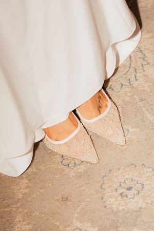 Close-up of bride wearing pearl-embellished pointed-toe flats beneath a flowing white wedding gown.