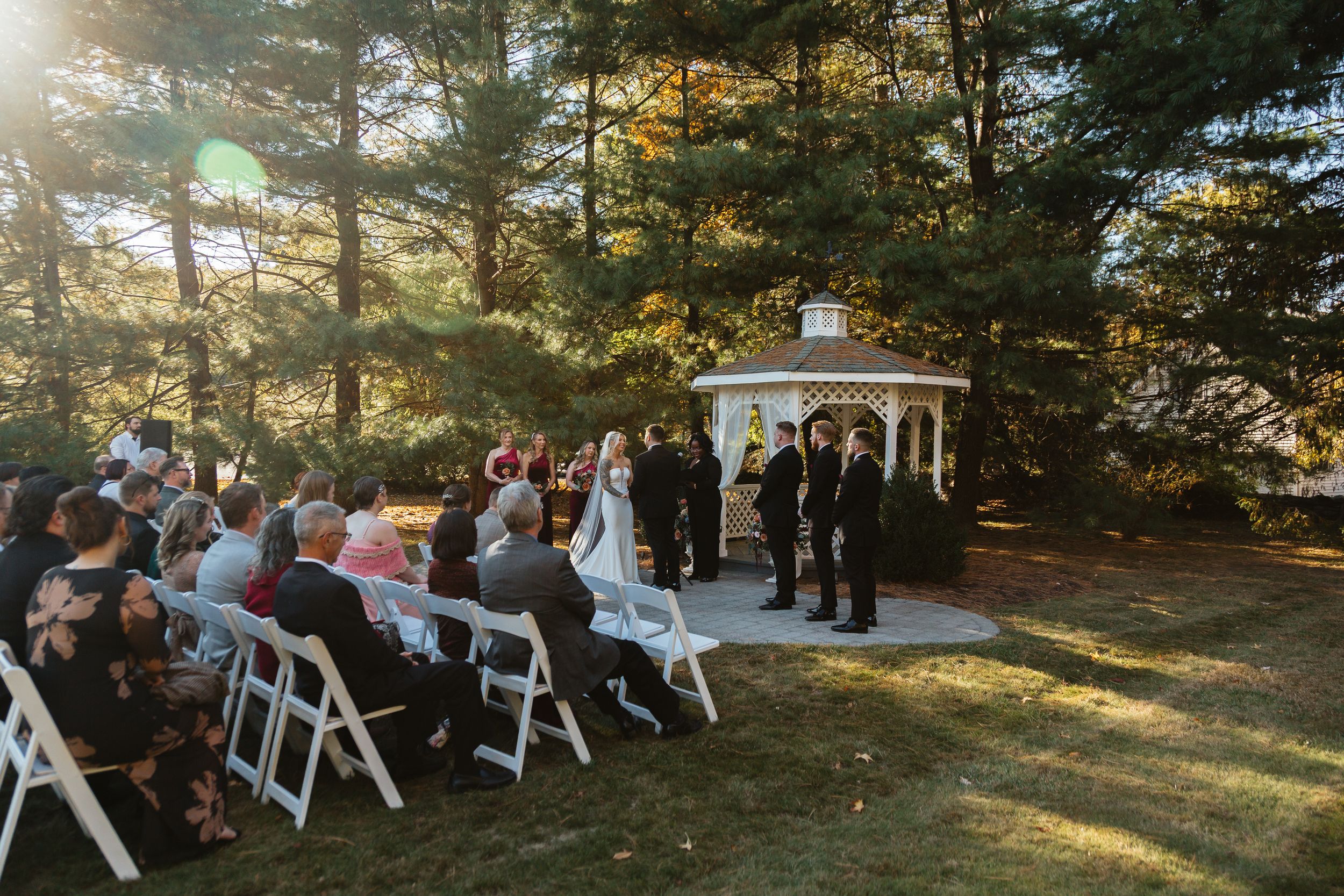 Outdoor wedding ceremony with guests seated in white chairs facing a white gazebo surrounded by tall pine trees.