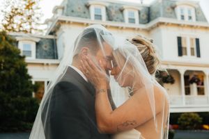 Bride and groom sharing an intimate moment under a flowing veil in front of a grand Victorian mansion.