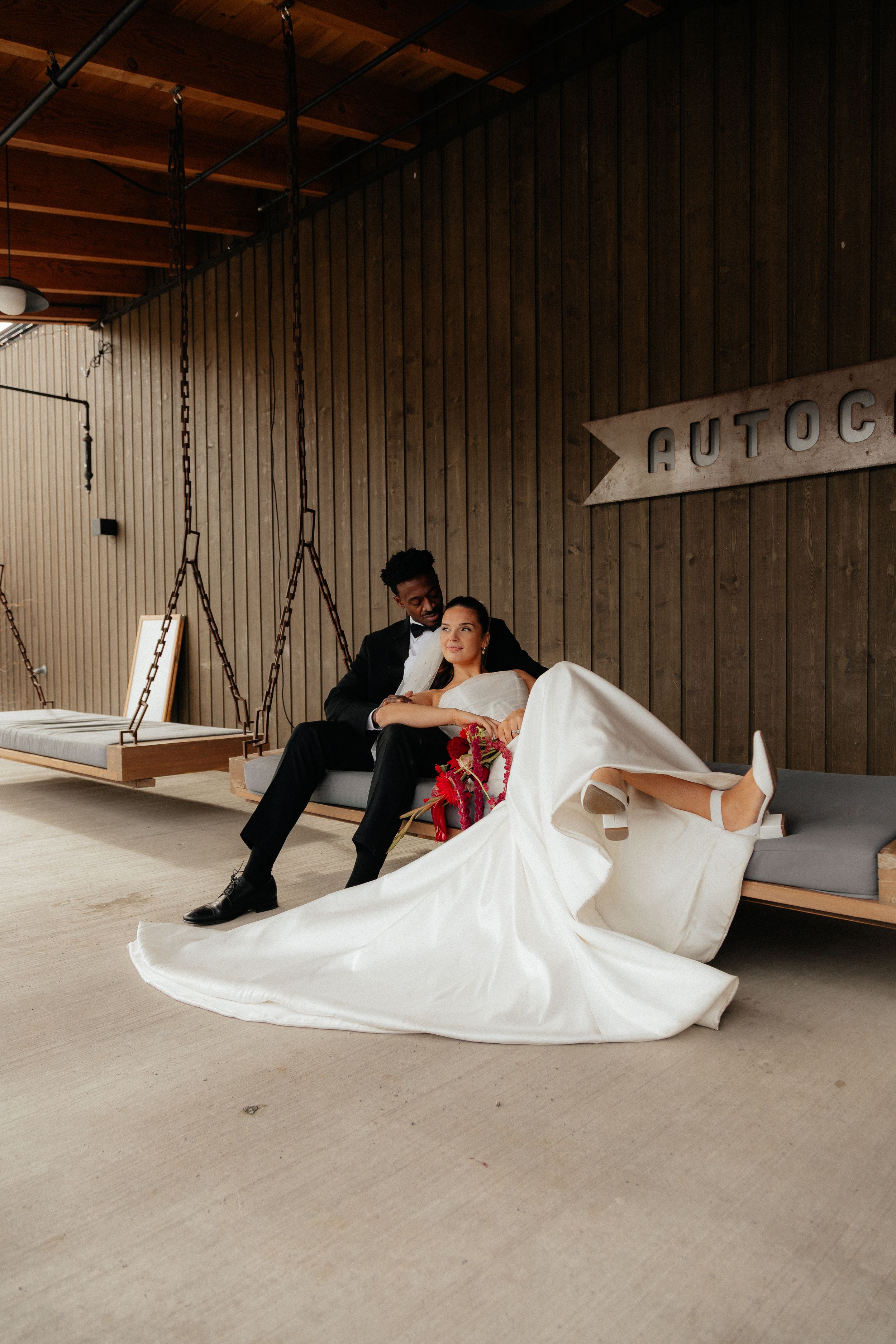 Bride and groom relaxing on hanging bench swings inside a modern rustic wooden venue space.
